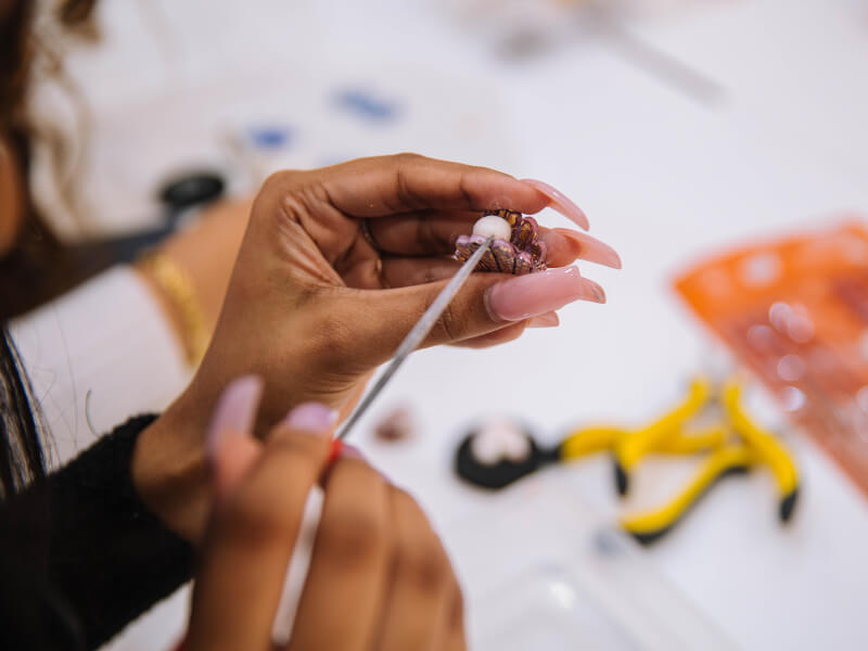Close up of hand with pink acrylic nails using a fine steel tool to insert a pearl into a clam DIY jewellery piece